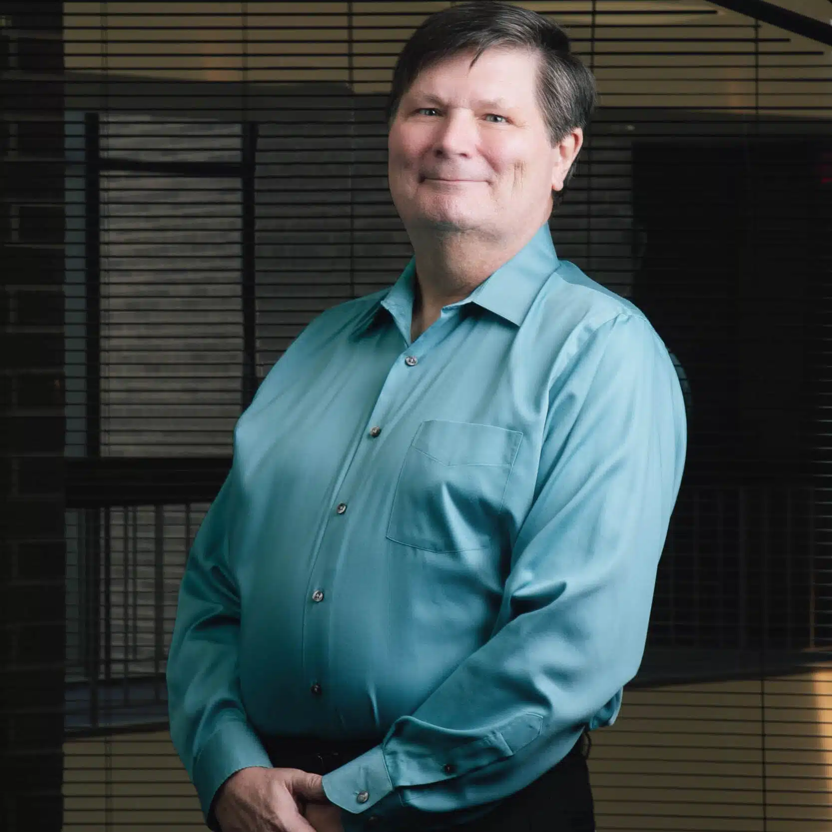 Smiling man with dark hair wearing a teal button-up shirt, standing with hands clasped in front of office window blinds.