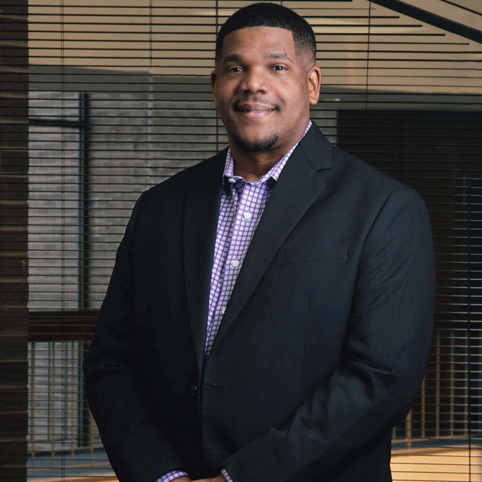 A smiling, short-haired man with hands casually clasped in front, wearing a purple checkered shirt under a black blazer.