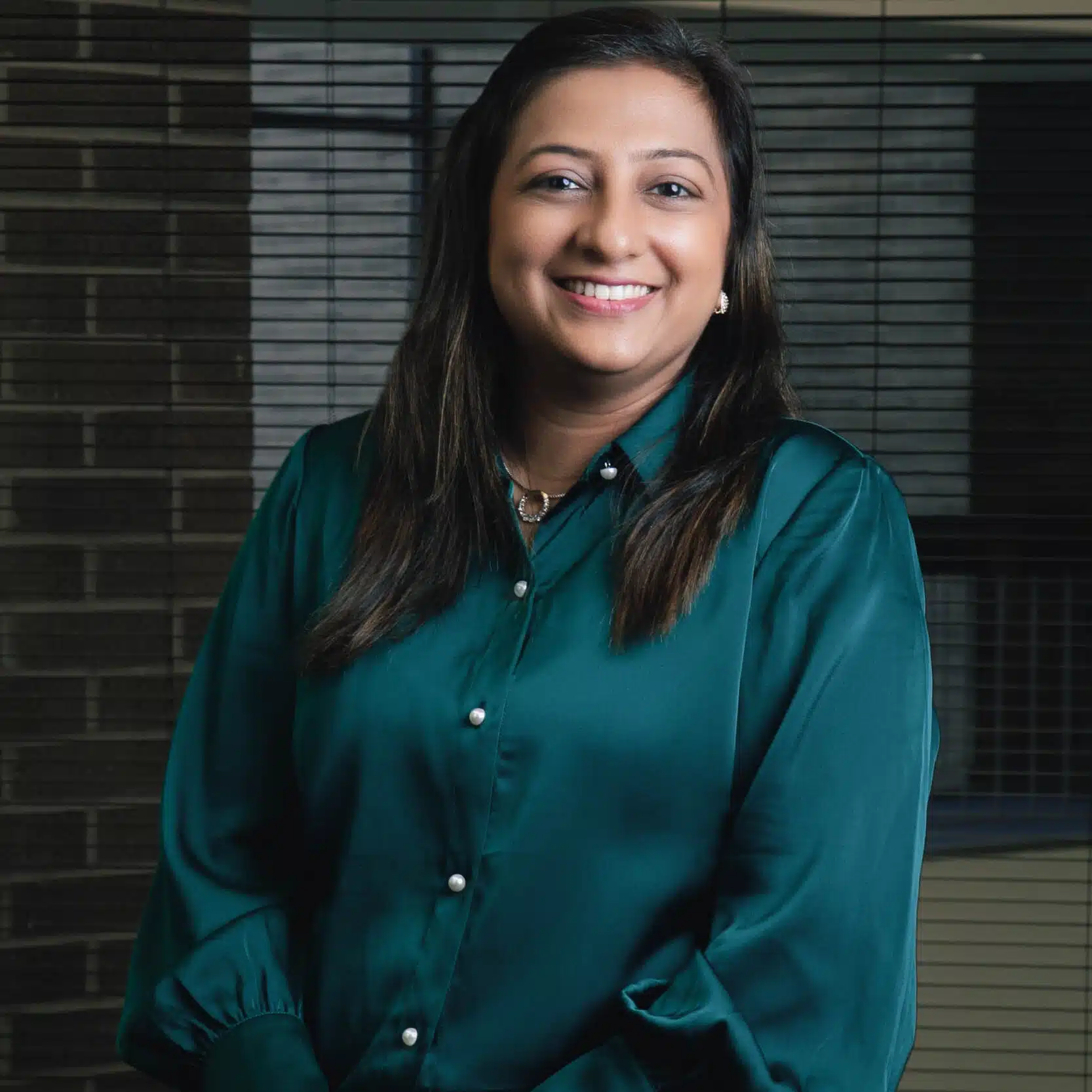 In an office setting, a smiling woman with long dark hair and a green satin blouse stands in front of window blinds.