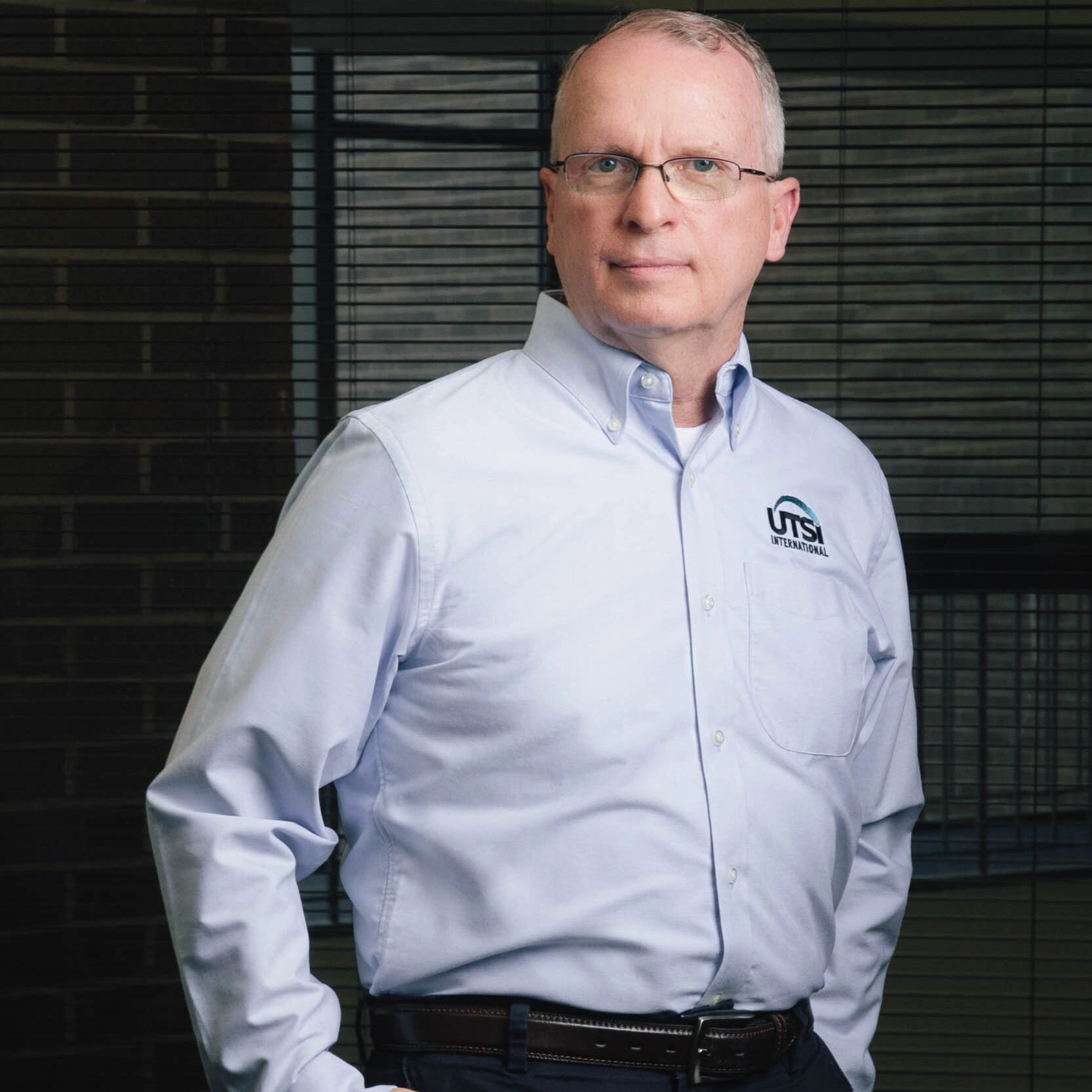 An image of a professional man standing tall and neutrally wearing glasses and a light blue UTSI International shirt.
