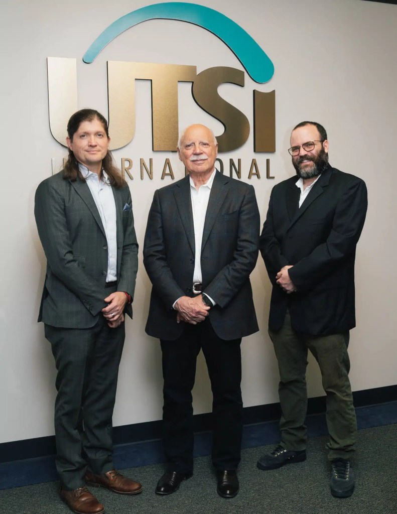 Posing formally for a corporate photo, three well-dressed men stand in front of a sizable UTSI International logo.