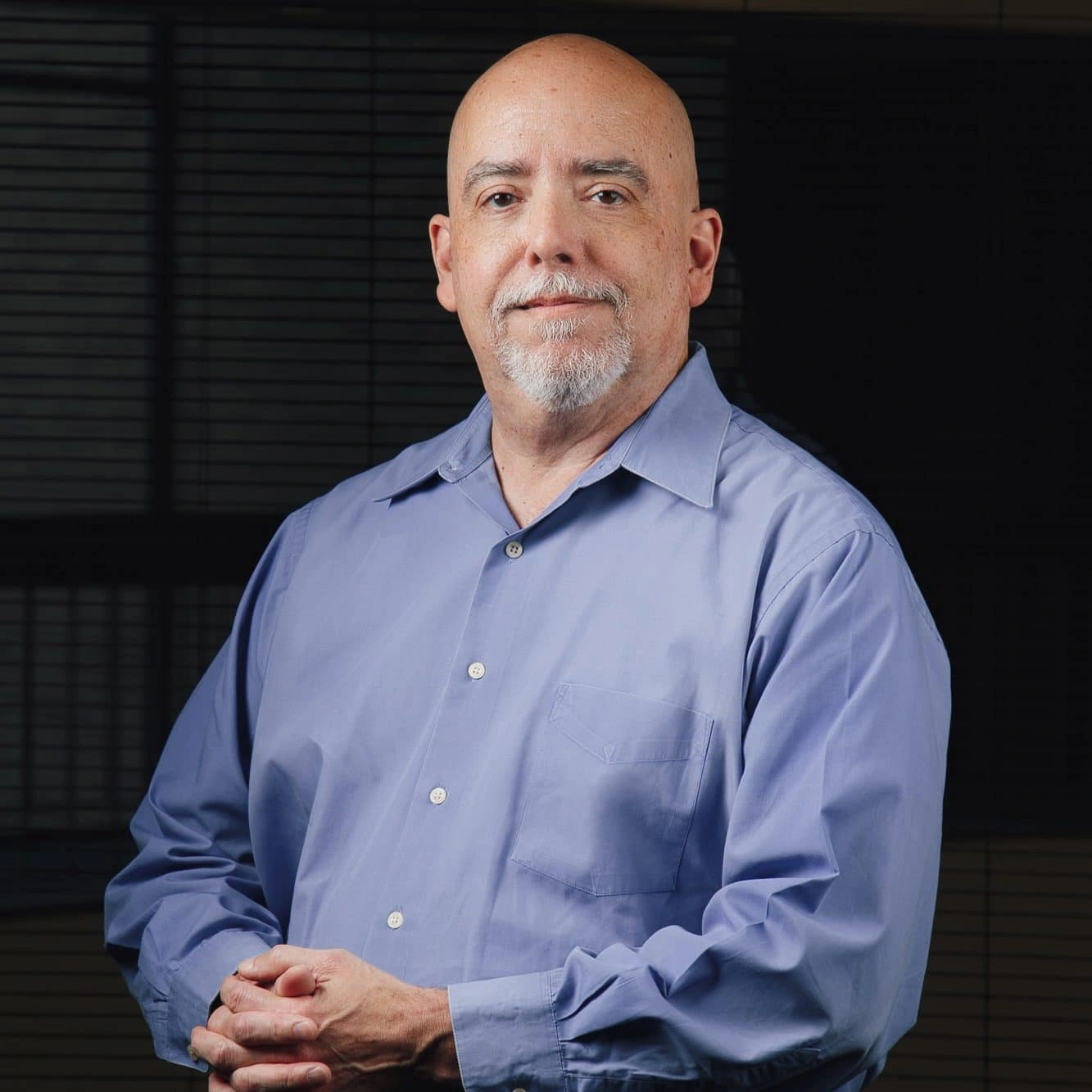 Standing calmly with his hands folded is a bald man with a gray beard, dressed in a light blue button-down shirt.